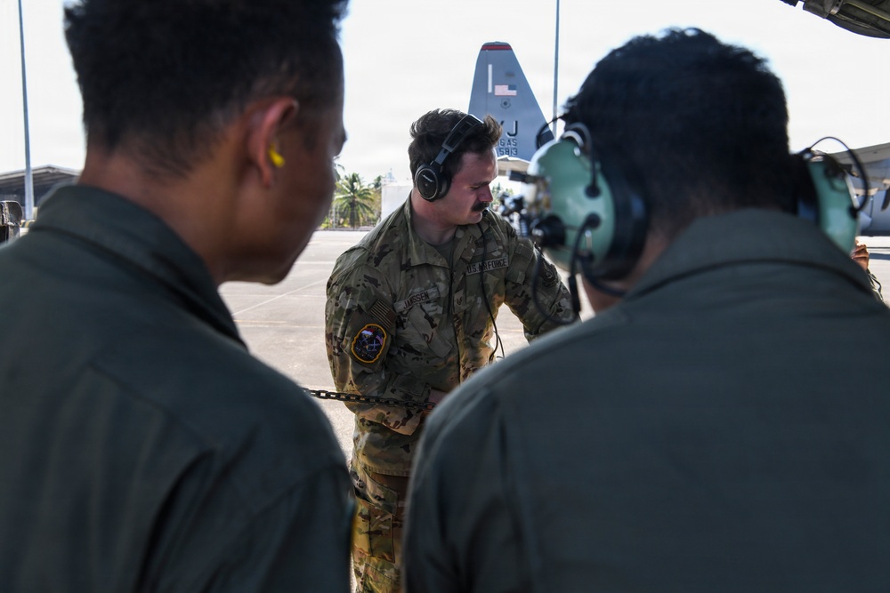 Three U.S. Air Force personnel are gathered on a flight line apron, appearing to conduct a briefing or pre-flight discussion. The central figure, wearing OCP (Operational Camouflage Pattern) uniform with a name tape reading 'JANSSE' and a U.S. Air Force patch, is wearing over-ear headphones and holding a chain. In the background, a military transport aircraft — identifiable as a C-130 by its tail fin with partial markings 'J' and numbers — is visible on the tarmac.