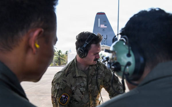 Three U.S. Air Force personnel are gathered on a flight line apron, appearing to conduct a briefing or pre-flight discussion. The central figure, wearing OCP (Operational Camouflage Pattern) uniform with a name tape reading 'JANSSE' and a U.S. Air Force patch, is wearing over-ear headphones and holding a chain. In the background, a military transport aircraft — identifiable as a C-130 by its tail fin with partial markings 'J' and numbers — is visible on the tarmac.
