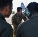 Three U.S. Air Force personnel are gathered on a flight line apron, appearing to conduct a briefing or pre-flight discussion. The central figure, wearing OCP (Operational Camouflage Pattern) uniform with a name tape reading 'JANSSE' and a U.S. Air Force patch, is wearing over-ear headphones and holding a chain. In the background, a military transport aircraft — identifiable as a C-130 by its tail fin with partial markings 'J' and numbers — is visible on the tarmac.