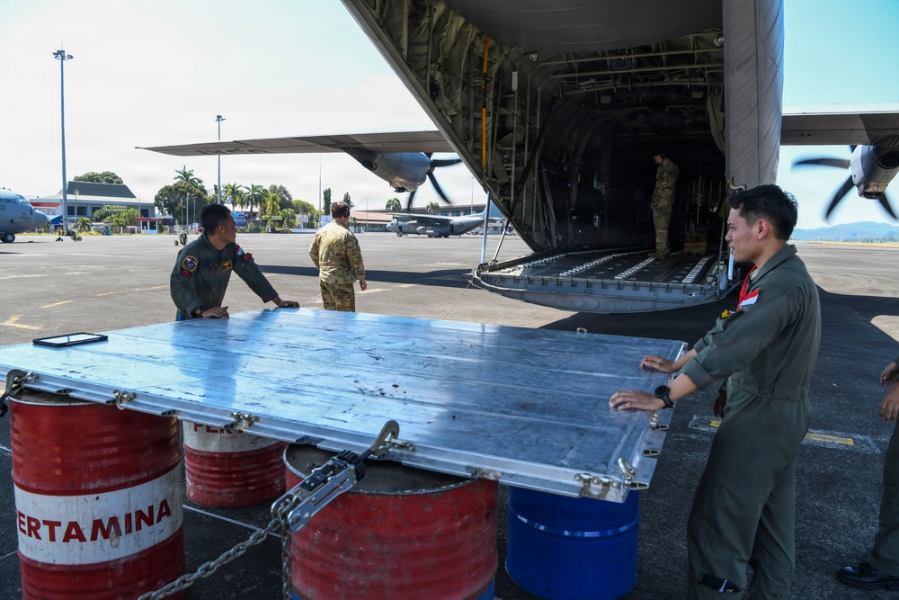 Military personnel are conducting cargo loading or unloading operations at an airfield, working around a large military transport aircraft with its rear ramp lowered. Red and blue Pertamina fuel drums are visible in the foreground, being used to support a metal platform or ramp extension. Personnel in olive green flight suits and one in a tan/multicam uniform are coordinating the ground operation.