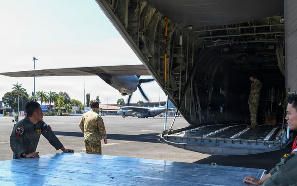 Military personnel are conducting cargo loading or unloading operations at an airfield, working around a large military transport aircraft with its rear ramp lowered. Red and blue Pertamina fuel drums are visible in the foreground, being used to support a metal platform or ramp extension. Personnel in olive green flight suits and one in a tan/multicam uniform are coordinating the ground operation.