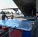 Military personnel are conducting cargo loading or unloading operations at an airfield, working around a large military transport aircraft with its rear ramp lowered. Red and blue Pertamina fuel drums are visible in the foreground, being used to support a metal platform or ramp extension. Personnel in olive green flight suits and one in a tan/multicam uniform are coordinating the ground operation.
