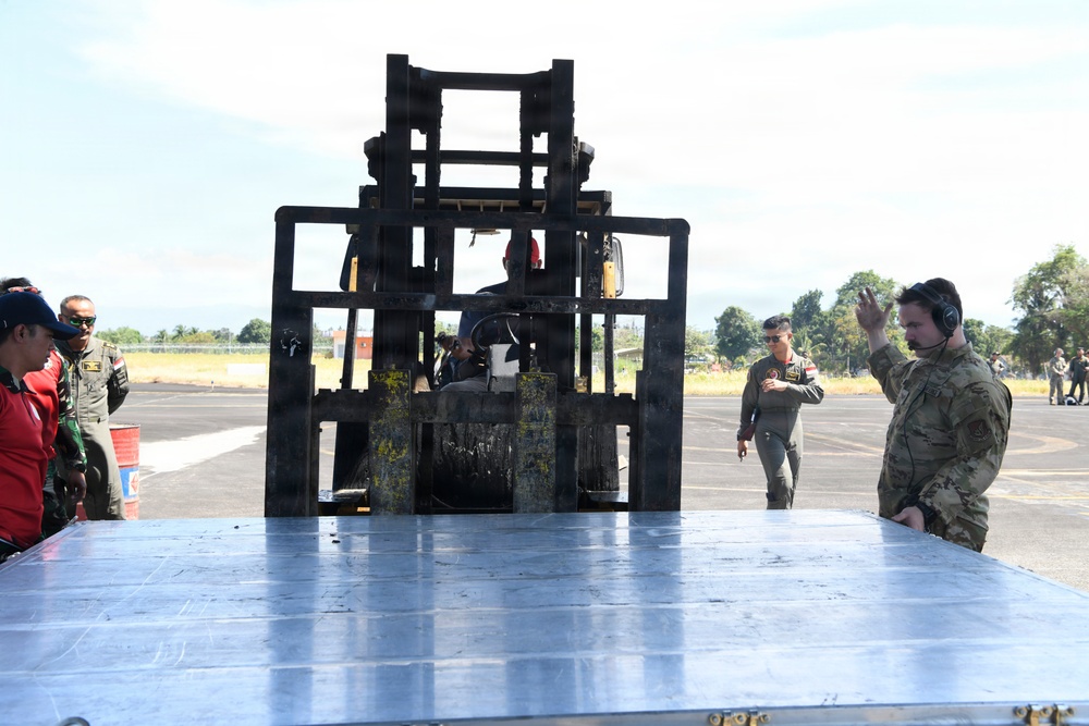 Military personnel are conducting a cargo loading or logistics operation on what appears to be an airfield tarmac. A forklift is positioned over a large metal pallet or cargo platform, with a soldier in multicam uniform giving hand signals to guide the operation. The scene involves a mixed group of personnel, suggesting a joint or multinational military exercise.