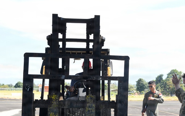 Military personnel are conducting a cargo loading or logistics operation on what appears to be an airfield tarmac. A forklift is positioned over a large metal pallet or cargo platform, with a soldier in multicam uniform giving hand signals to guide the operation. The scene involves a mixed group of personnel, suggesting a joint or multinational military exercise.