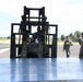 Military personnel are conducting a cargo loading or logistics operation on what appears to be an airfield tarmac. A forklift is positioned over a large metal pallet or cargo platform, with a soldier in multicam uniform giving hand signals to guide the operation. The scene involves a mixed group of personnel, suggesting a joint or multinational military exercise.