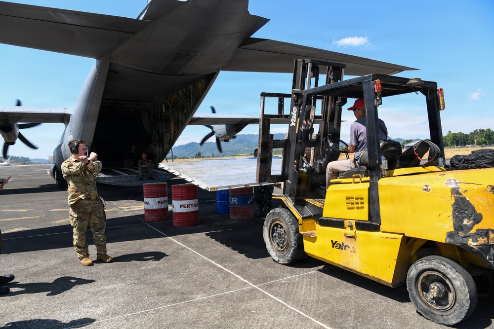 A military cargo aircraft, likely a C-130 Hercules, is being loaded or unloaded on an airfield tarmac. A soldier in camouflage uniform with hearing protection is directing a Yale forklift operator who is maneuvering cargo toward the open rear ramp of the aircraft. Red and blue fuel barrels marked 'PERTAMINA' are visible in the background, suggesting this operation is taking place in Indonesia.