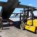 A military cargo aircraft, likely a C-130 Hercules, is being loaded or unloaded on an airfield tarmac. A soldier in camouflage uniform with hearing protection is directing a Yale forklift operator who is maneuvering cargo toward the open rear ramp of the aircraft. Red and blue fuel barrels marked 'PERTAMINA' are visible in the background, suggesting this operation is taking place in Indonesia.