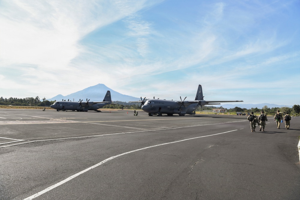 Two U.S. Air Force C-130 Hercules transport aircraft are parked on a tarmac at what appears to be a military airfield in a tropical or mountainous region, with a prominent volcanic peak visible in the background. Several military personnel in combat gear are walking across the tarmac toward the aircraft. The setting suggests a forward operating location or joint military exercise.