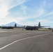 Two U.S. Air Force C-130 Hercules transport aircraft are parked on a tarmac at what appears to be a military airfield in a tropical or mountainous region, with a prominent volcanic peak visible in the background. Several military personnel in combat gear are walking across the tarmac toward the aircraft. The setting suggests a forward operating location or joint military exercise.