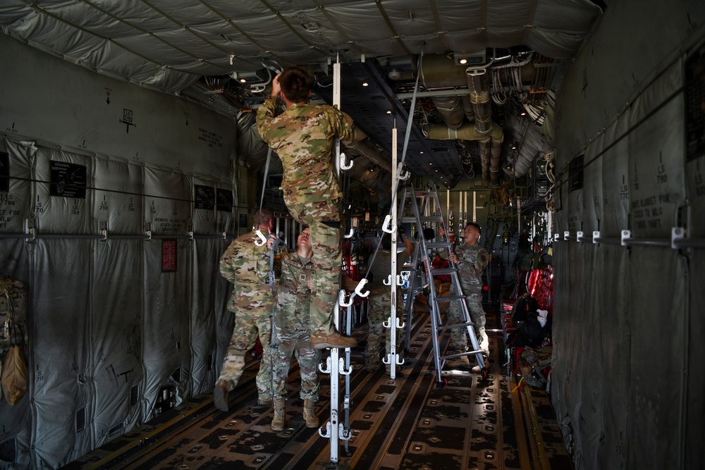 Military personnel in camouflage uniforms are working inside the cargo hold of a large military transport aircraft, likely a C-130 or C-17. One soldier is climbing a ladder to perform maintenance or installation work on the overhead systems, while others assist from below. The interior shows typical military cargo aircraft features including tie-down rings, padded sidewalls, and overhead ducting.
