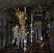 Military personnel in camouflage uniforms are working inside the cargo hold of a large military transport aircraft, likely a C-130 or C-17. One soldier is climbing a ladder to perform maintenance or installation work on the overhead systems, while others assist from below. The interior shows typical military cargo aircraft features including tie-down rings, padded sidewalls, and overhead ducting.