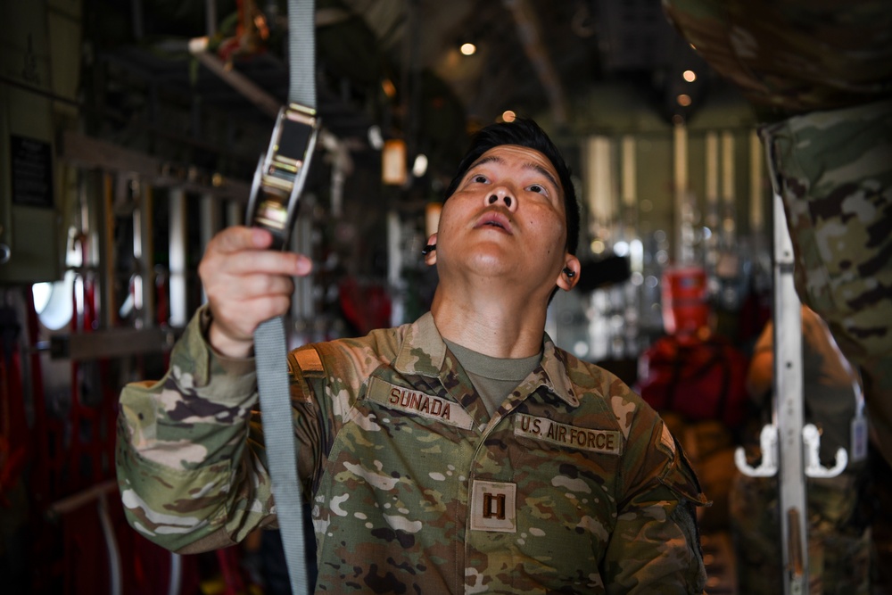 A U.S. Air Force Captain named Sunada is working inside what appears to be a military transport aircraft, inspecting or adjusting a cargo strap or tie-down mechanism. He is wearing OCP (Operational Camouflage Pattern) uniform with a Captain's rank insignia and is looking upward while handling the strap. The interior of the aircraft shows typical cargo hold features including overhead lighting, tie-down rails, and equipment.