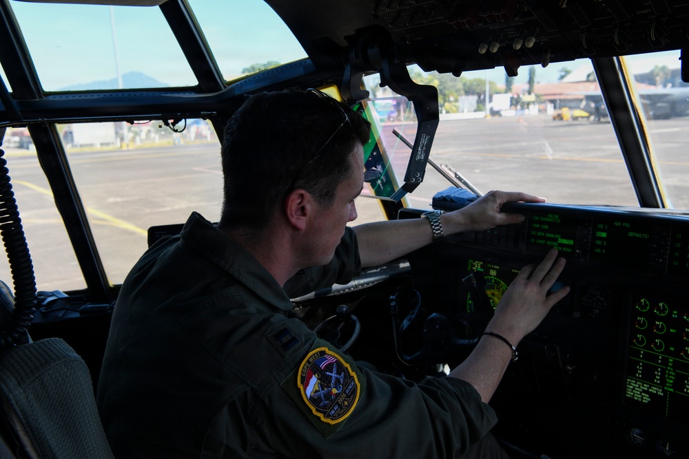 A military aviator in an olive drab flight suit is seated in the cockpit of a large military aircraft, operating or inspecting the instrument panel. The pilot wears a mission patch on his left shoulder and has sunglasses resting on his head. The aircraft appears to be parked on a flight line or tarmac, visible through the large cockpit windows.