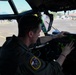 A military aviator in an olive drab flight suit is seated in the cockpit of a large military aircraft, operating or inspecting the instrument panel. The pilot wears a mission patch on his left shoulder and has sunglasses resting on his head. The aircraft appears to be parked on a flight line or tarmac, visible through the large cockpit windows.