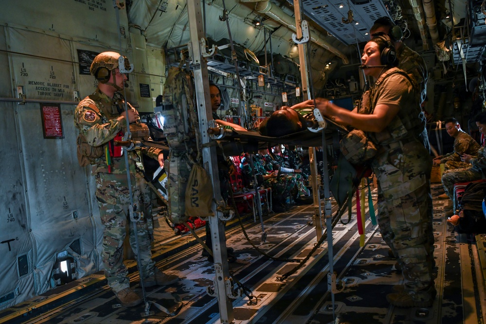 Military medical personnel are conducting an aeromedical evacuation inside what appears to be a C-130 military transport aircraft. At least one patient is secured on a litter/stretcher in a stacked litter system, being attended to by uniformed personnel wearing combat helmets and headsets. Additional personnel and patients are visible further back in the aircraft cabin.