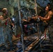 Military medical personnel are conducting an aeromedical evacuation inside what appears to be a C-130 military transport aircraft. At least one patient is secured on a litter/stretcher in a stacked litter system, being attended to by uniformed personnel wearing combat helmets and headsets. Additional personnel and patients are visible further back in the aircraft cabin.
