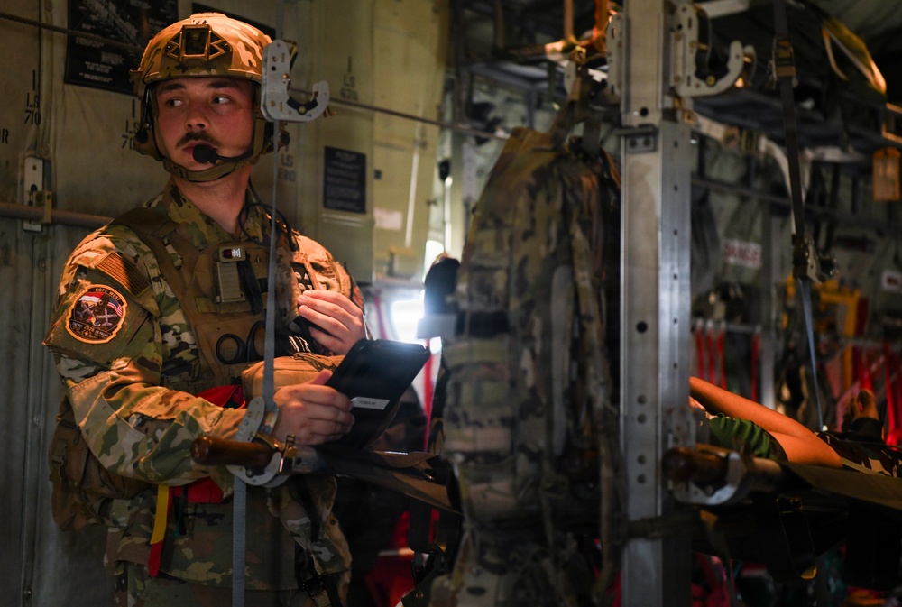 A military service member in full combat gear stands inside what appears to be a military transport aircraft, likely a C-130 or similar cargo plane. The individual is wearing a tactical helmet with communications equipment and a microphone, holding an illuminated device or tablet. The interior of the aircraft shows cargo straps, litter/stretcher equipment, and structural framing consistent with an aeromedical evacuation or airborne operations configuration.