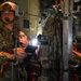 A military service member in full combat gear stands inside what appears to be a military transport aircraft, likely a C-130 or similar cargo plane. The individual is wearing a tactical helmet with communications equipment and a microphone, holding an illuminated device or tablet. The interior of the aircraft shows cargo straps, litter/stretcher equipment, and structural framing consistent with an aeromedical evacuation or airborne operations configuration.