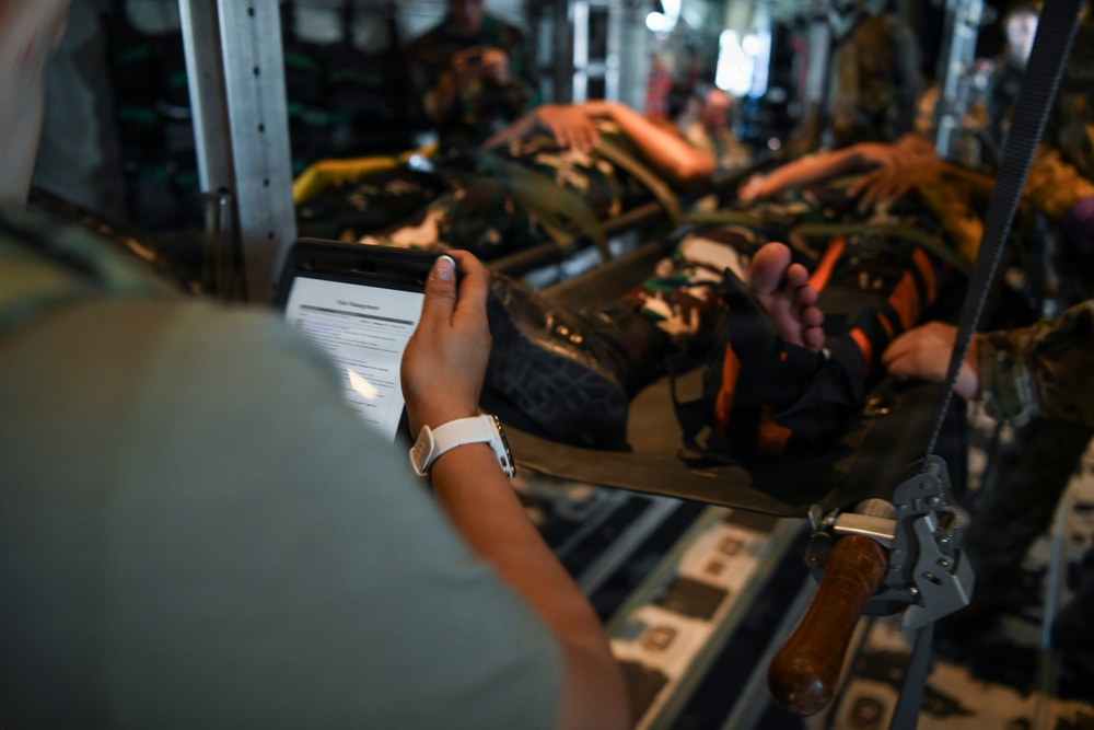 The image shows what appears to be a military aeromedical evacuation scenario inside an aircraft cargo hold. A person in military uniform is holding a tablet device displaying a document, while patients on litters (stretchers) are visible in the background secured to the aircraft's interior walls. The setting and equipment suggest a medical training exercise or actual casualty evacuation operation.