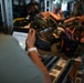 The image shows what appears to be a military aeromedical evacuation scenario inside an aircraft cargo hold. A person in military uniform is holding a tablet device displaying a document, while patients on litters (stretchers) are visible in the background secured to the aircraft's interior walls. The setting and equipment suggest a medical training exercise or actual casualty evacuation operation.