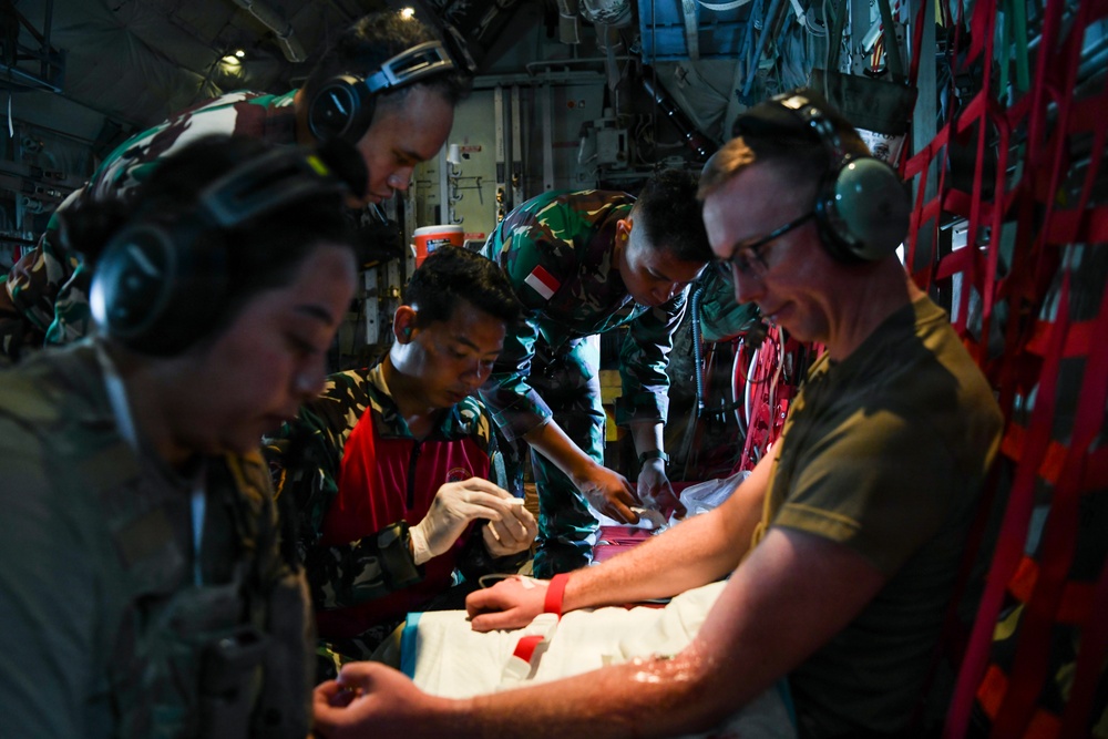 Military medical personnel are performing what appears to be an aeromedical evacuation or medical treatment inside the cargo hold of a military aircraft. The team includes personnel wearing Indonesian military camouflage uniforms (identifiable by the red-and-white Indonesian flag patch) alongside what appears to be a Western military service member in an olive t-shirt. They are collectively attending to a patient on a stretcher or litter.