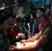 Military medical personnel are performing what appears to be an aeromedical evacuation or medical treatment inside the cargo hold of a military aircraft. The team includes personnel wearing Indonesian military camouflage uniforms (identifiable by the red-and-white Indonesian flag patch) alongside what appears to be a Western military service member in an olive t-shirt. They are collectively attending to a patient on a stretcher or litter.