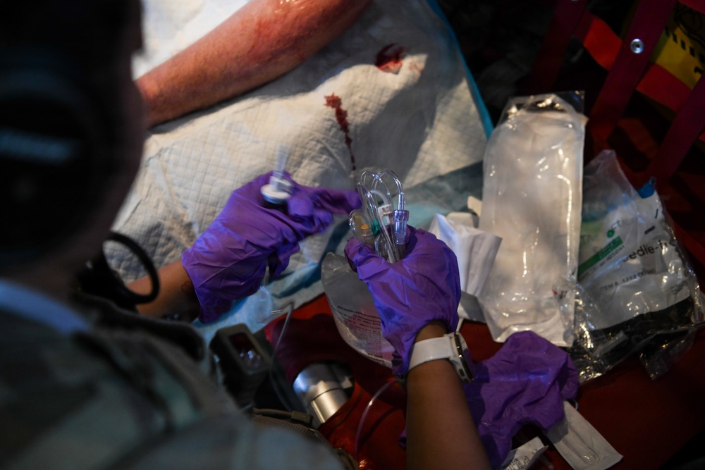 A medical professional wearing purple nitrile gloves is preparing or administering intravenous (IV) equipment in what appears to be an emergency or field medical setting. Visible blood on absorbent padding and multiple opened medical supply packages suggest active trauma care is being performed. The scene indicates urgent pre-hospital or tactical medical treatment.