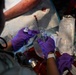 A medical professional wearing purple nitrile gloves is preparing or administering intravenous (IV) equipment in what appears to be an emergency or field medical setting. Visible blood on absorbent padding and multiple opened medical supply packages suggest active trauma care is being performed. The scene indicates urgent pre-hospital or tactical medical treatment.