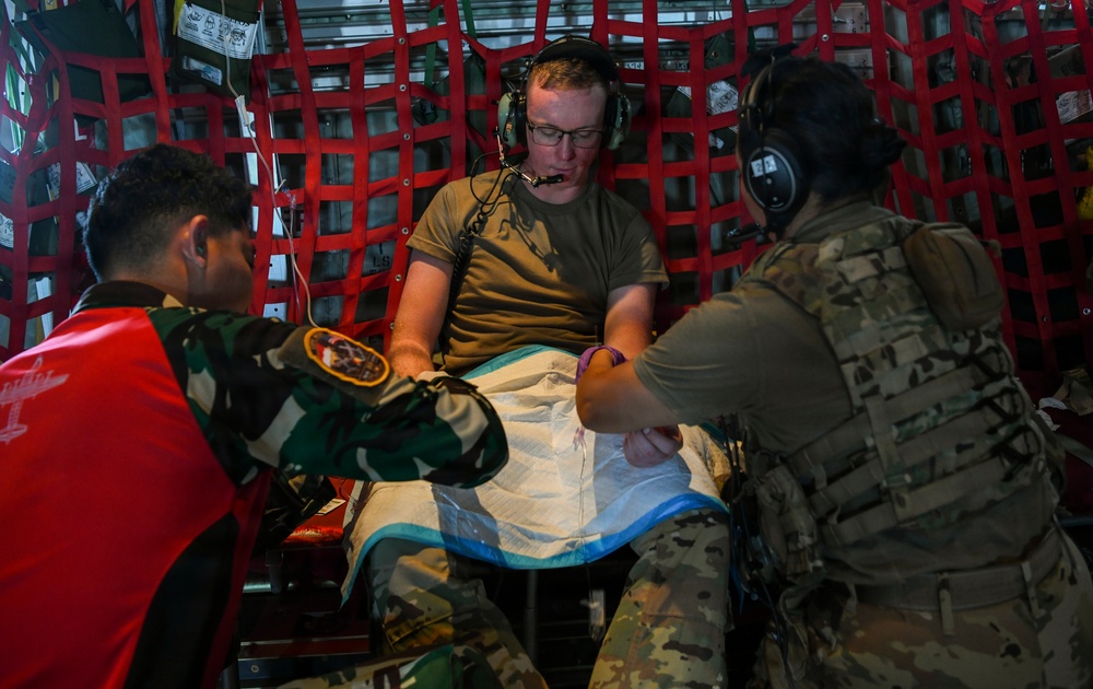 Three individuals are inside what appears to be a military aircraft, engaged in a medical training or treatment scenario. A person in an olive t-shirt wearing aviation headsets is seated as a simulated patient, while two others in military uniforms — one in camouflage with a red vest and one in multicam — attend to his lower body area covered with a drape. The setting suggests a joint military medical exercise or aeromedical evacuation training.