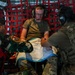 Three individuals are inside what appears to be a military aircraft, engaged in a medical training or treatment scenario. A person in an olive t-shirt wearing aviation headsets is seated as a simulated patient, while two others in military uniforms — one in camouflage with a red vest and one in multicam — attend to his lower body area covered with a drape. The setting suggests a joint military medical exercise or aeromedical evacuation training.