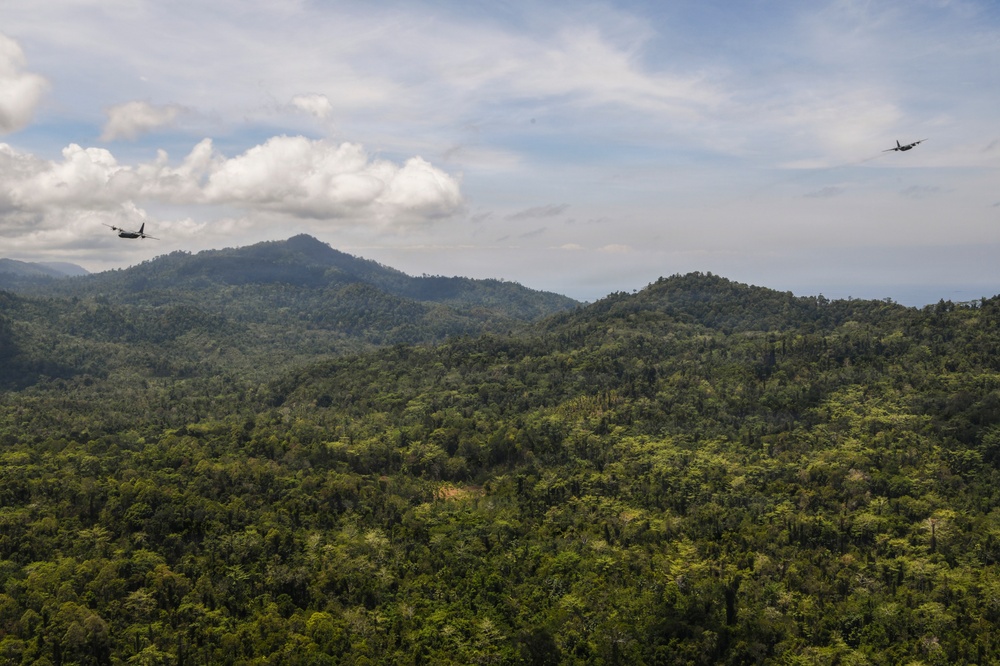 An aerial view of a dense tropical rainforest covering rolling hills and mountains under a partly cloudy sky. Two military-style aircraft are visible in flight over the forested landscape, one on the left and one on the right side of the frame. A small patch of exposed reddish-brown earth is visible amid the otherwise unbroken green canopy.