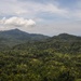 An aerial view of a dense tropical rainforest covering rolling hills and mountains under a partly cloudy sky. Two military-style aircraft are visible in flight over the forested landscape, one on the left and one on the right side of the frame. A small patch of exposed reddish-brown earth is visible amid the otherwise unbroken green canopy.