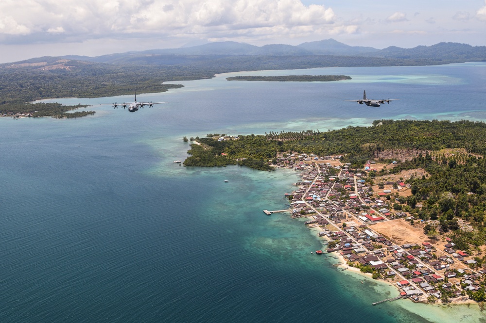 An aerial photograph shows two military turboprop transport aircraft flying in formation over a tropical coastal island settlement. The image captures a small seaside town with dense vegetation, shallow turquoise reef waters, and larger forested islands visible in the background. The aircraft appear to be on a low-altitude pass or patrol mission over the archipelago.