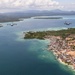 An aerial photograph shows two military turboprop transport aircraft flying in formation over a tropical coastal island settlement. The image captures a small seaside town with dense vegetation, shallow turquoise reef waters, and larger forested islands visible in the background. The aircraft appear to be on a low-altitude pass or patrol mission over the archipelago.