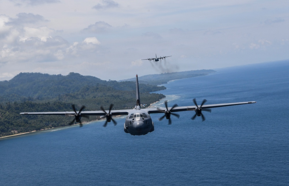 Two military turboprop transport aircraft, identifiable as C-130 Hercules variants, are flying in formation over a coastal tropical region. The foreground aircraft is captured in sharp detail showing all four spinning propeller engines, while a second aircraft is visible in the background trailing light exhaust smoke. The terrain below features a densely forested coastline with blue ocean waters and a sandy beach shoreline.