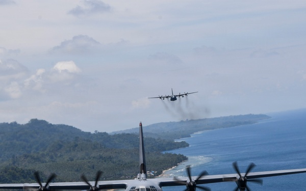 Two military turboprop transport aircraft, identifiable as C-130 Hercules variants, are flying in formation over a coastal tropical region. The foreground aircraft is captured in sharp detail showing all four spinning propeller engines, while a second aircraft is visible in the background trailing light exhaust smoke. The terrain below features a densely forested coastline with blue ocean waters and a sandy beach shoreline.