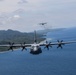 Two military turboprop transport aircraft, identifiable as C-130 Hercules variants, are flying in formation over a coastal tropical region. The foreground aircraft is captured in sharp detail showing all four spinning propeller engines, while a second aircraft is visible in the background trailing light exhaust smoke. The terrain below features a densely forested coastline with blue ocean waters and a sandy beach shoreline.