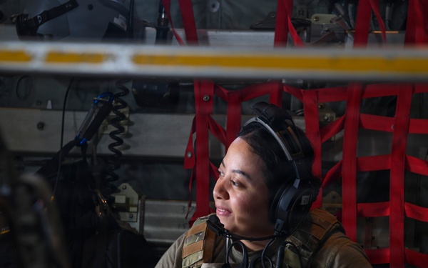 A female military service member is seated inside what appears to be a military transport aircraft, wearing tactical gear and aviation headphones. She is looking to her left with a slight smile, holding an object in her hands. The interior of the aircraft is visible behind her, including red cargo netting and various equipment panels.