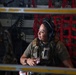 A female military service member is seated inside what appears to be a military transport aircraft, wearing tactical gear and aviation headphones. She is looking to her left with a slight smile, holding an object in her hands. The interior of the aircraft is visible behind her, including red cargo netting and various equipment panels.