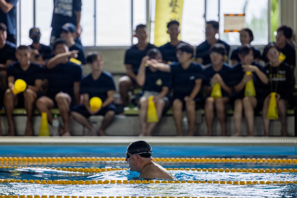 A swimmer wearing a black cap and goggles is in an indoor swimming pool, observed by a large group of spectators seated in the background. The spectators are uniformly dressed in dark navy/black shirts, suggesting an organized team or group event. Yellow lane dividers are visible in the pool, and yellow accessories and banners are present throughout the scene.