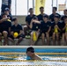 A swimmer wearing a black cap and goggles is in an indoor swimming pool, observed by a large group of spectators seated in the background. The spectators are uniformly dressed in dark navy/black shirts, suggesting an organized team or group event. Yellow lane dividers are visible in the pool, and yellow accessories and banners are present throughout the scene.