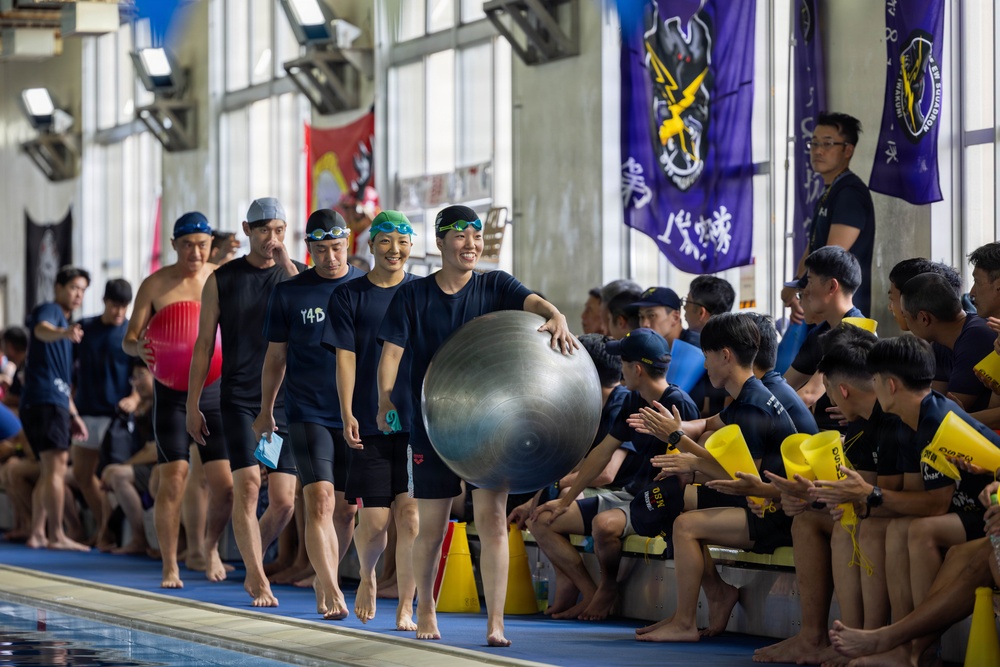 A group of swimmers in swim caps and dark athletic wear walk along the edge of an indoor swimming pool, with one participant carrying a large silver exercise ball. They are greeted by a row of seated spectators or teammates holding yellow clappers/noisemakers. The setting appears to be an organized aquatic competition or team event, with decorative flags and banners hanging in the background.