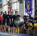 A group of swimmers in swim caps and dark athletic wear walk along the edge of an indoor swimming pool, with one participant carrying a large silver exercise ball. They are greeted by a row of seated spectators or teammates holding yellow clappers/noisemakers. The setting appears to be an organized aquatic competition or team event, with decorative flags and banners hanging in the background.