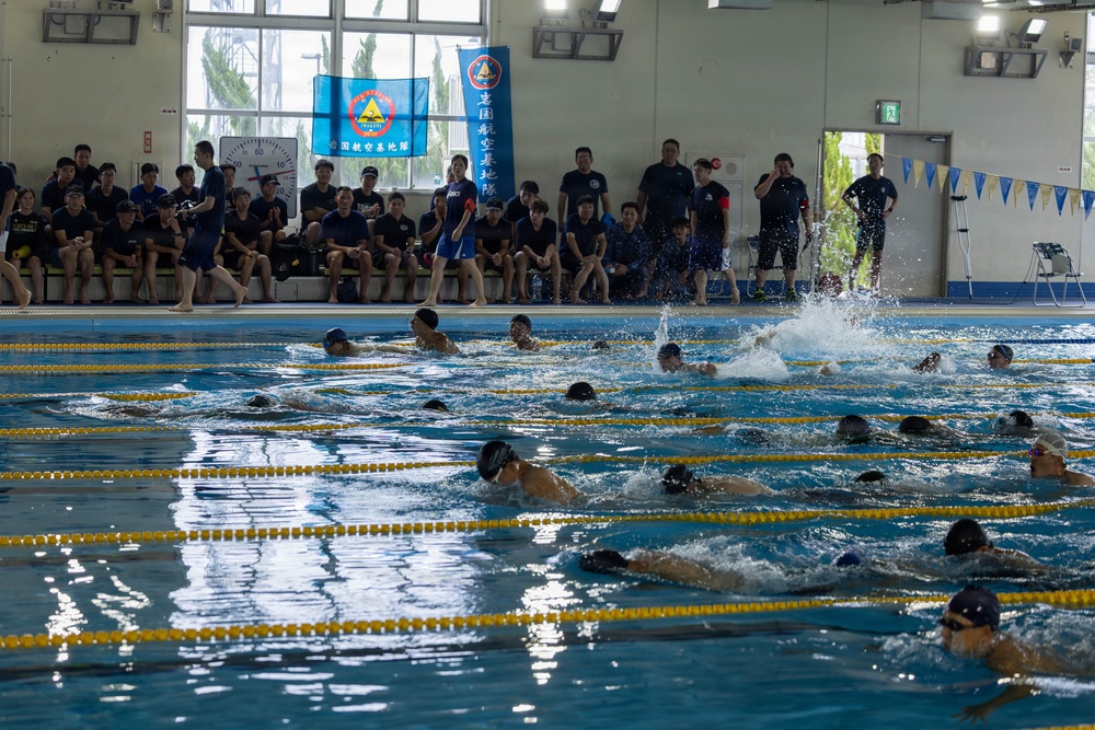 An indoor swimming competition or training event is taking place at a pool facility with Japanese signage. Multiple swimmers are actively swimming in lane-divided lanes while a large group of spectators and officials observe from the poolside. Banners with Japanese text and organizational emblems are displayed on the wall in the background.