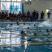 An indoor swimming competition or training event is taking place at a pool facility with Japanese signage. Multiple swimmers are actively swimming in lane-divided lanes while a large group of spectators and officials observe from the poolside. Banners with Japanese text and organizational emblems are displayed on the wall in the background.