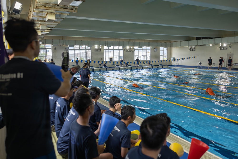 An indoor swimming pool event is underway, with swimmers in orange gear actively racing or training in the lanes. A large group of spectators and participants wearing matching navy blue shirts are gathered along the poolside, watching and recording the activity. The setting appears to be an organized aquatic training or competition event.