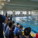 An indoor swimming pool event is underway, with swimmers in orange gear actively racing or training in the lanes. A large group of spectators and participants wearing matching navy blue shirts are gathered along the poolside, watching and recording the activity. The setting appears to be an organized aquatic training or competition event.