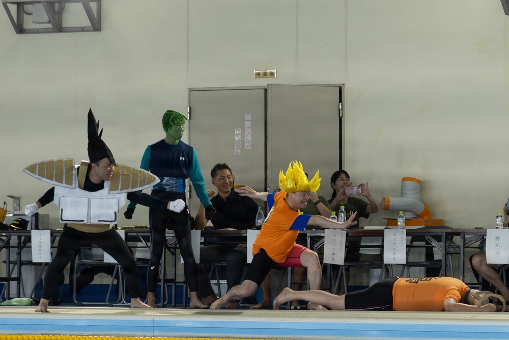 A cosplay or costume performance is taking place at an indoor swimming pool facility, likely in Japan based on the Japanese text visible on signs. Participants are dressed in various costumes including a character with a yellow spiky wig resembling a Super Saiyan from Dragon Ball Z, a person with green face paint, and someone wearing a large flat disc/hat costume. Spectators and officials are seated at tables along the poolside watching and photographing the event.