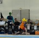 A cosplay or costume performance is taking place at an indoor swimming pool facility, likely in Japan based on the Japanese text visible on signs. Participants are dressed in various costumes including a character with a yellow spiky wig resembling a Super Saiyan from Dragon Ball Z, a person with green face paint, and someone wearing a large flat disc/hat costume. Spectators and officials are seated at tables along the poolside watching and photographing the event.