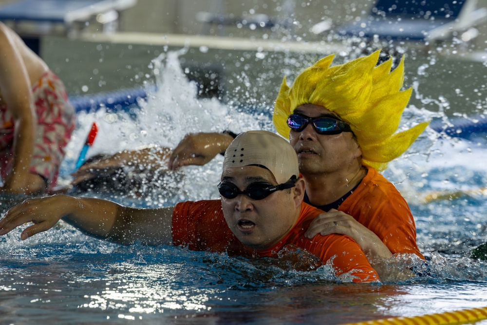 Two individuals are in a swimming pool, both wearing orange shirts and swim goggles. The person in the background is wearing a distinctive bright yellow spiky wig resembling an anime character (likely inspired by Dragon Ball Z's Super Saiyan style). The foreground person wears a beige swim cap, and both appear to be participating in an assisted or recreational swimming activity.